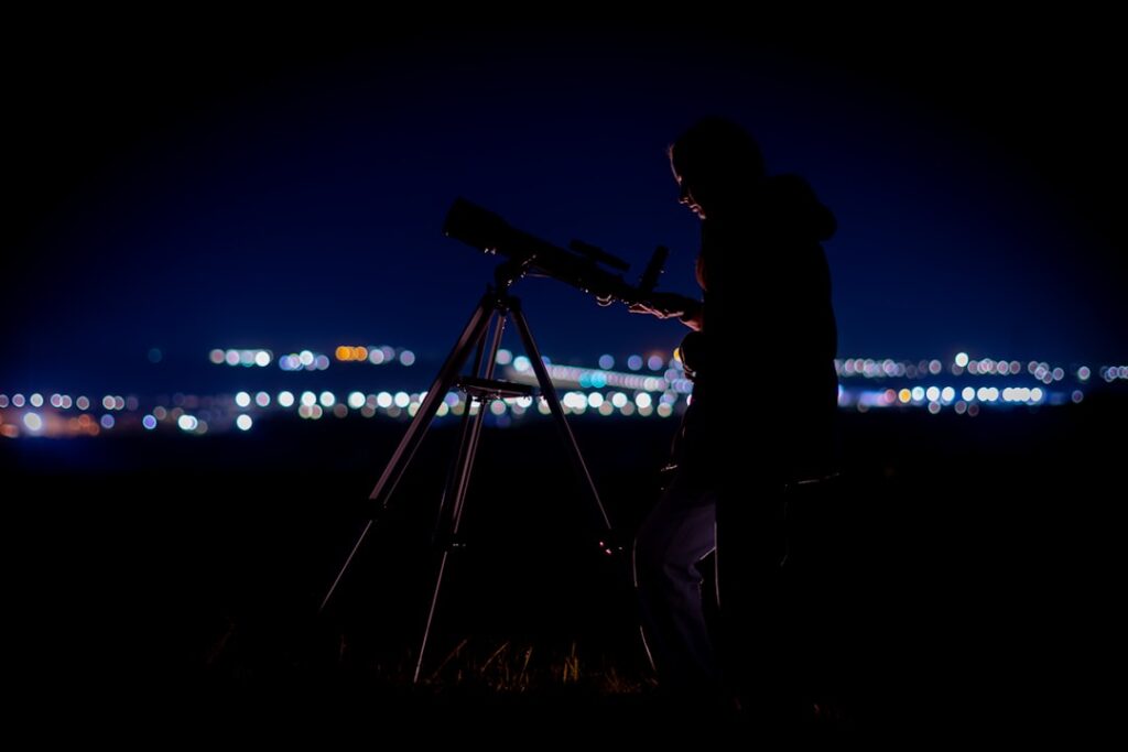 Person looking through a telescope at night with city lights.
