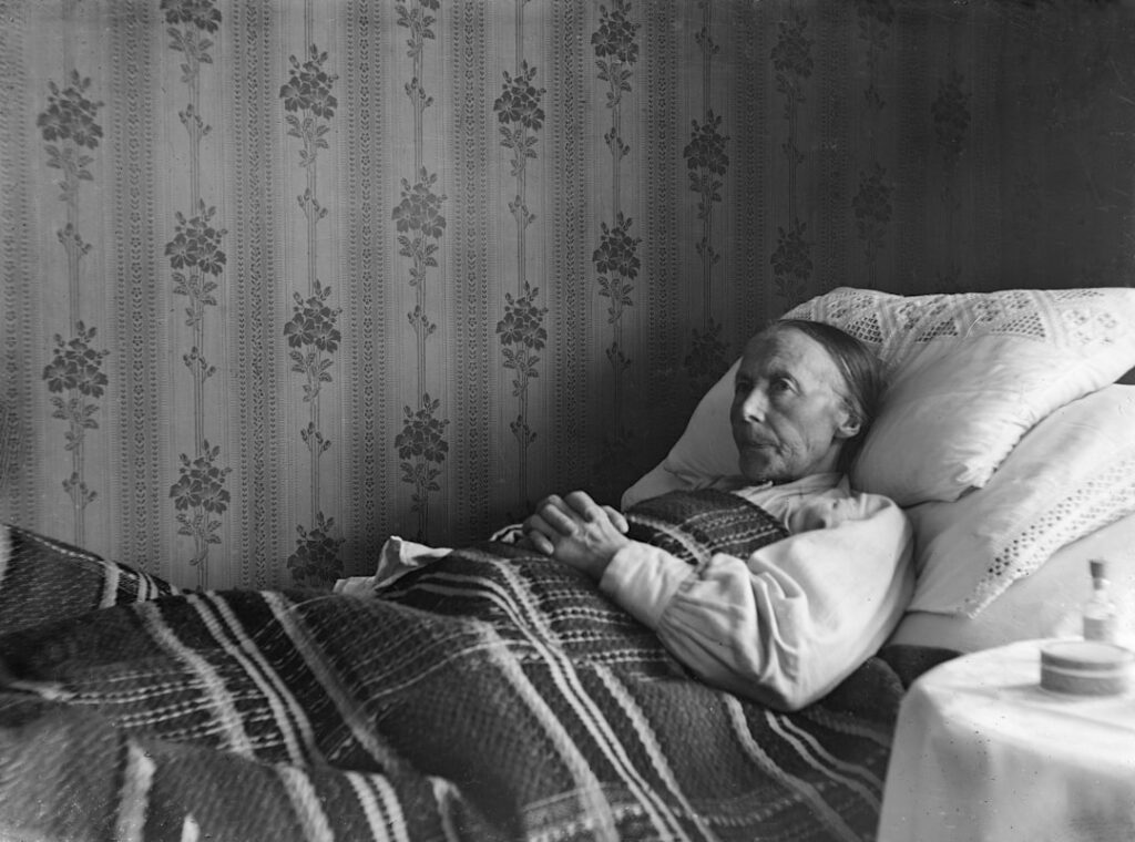 An elderly person lies in bed, covered by a blanket.