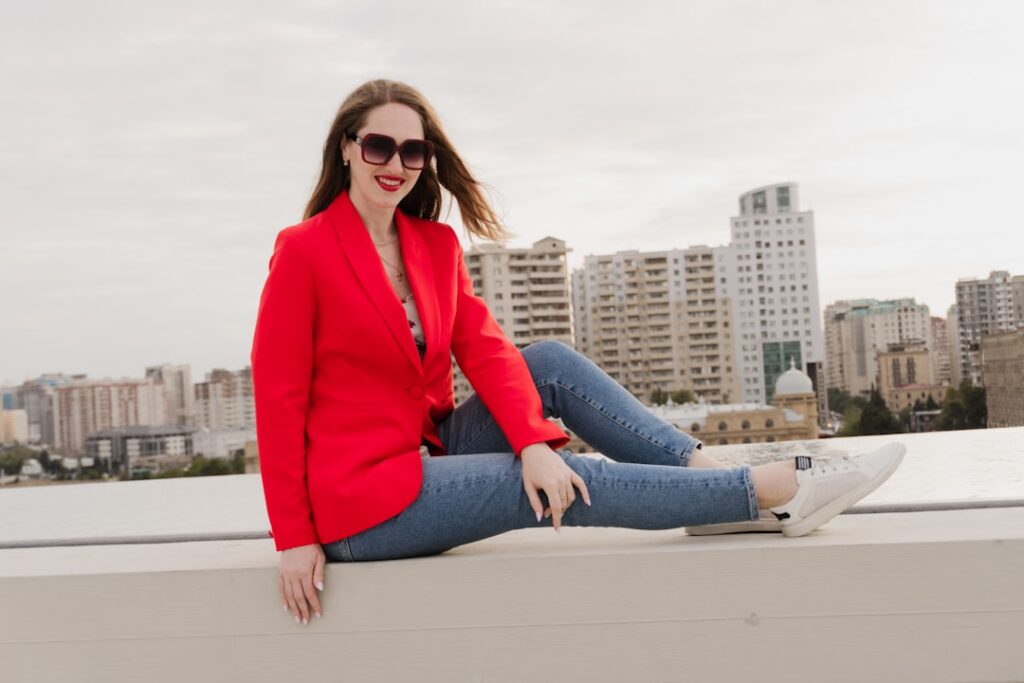 Woman in red blazer and jeans sits outdoors.