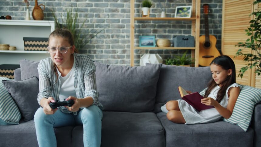 Woman plays video game while girl reads book on couch.