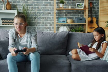 Woman plays video game while girl reads book on couch.