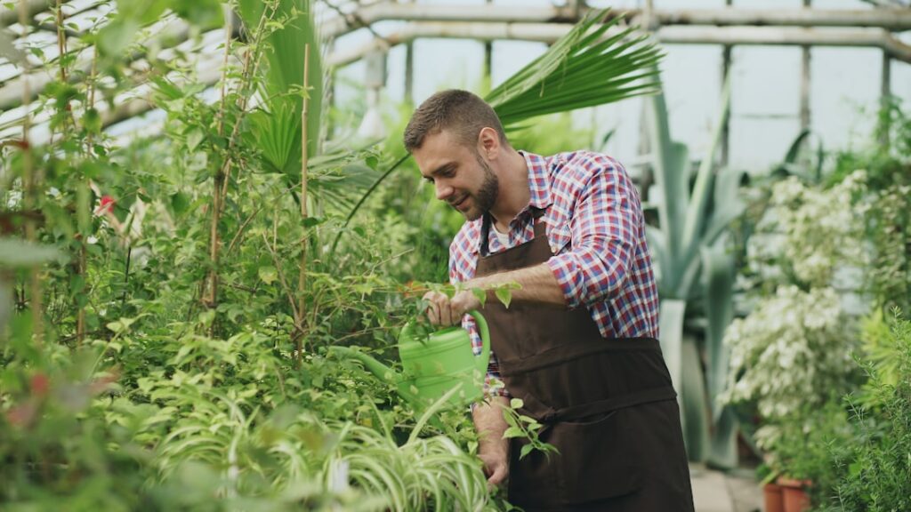 Man watering plants in a greenhouse with lush greenery.