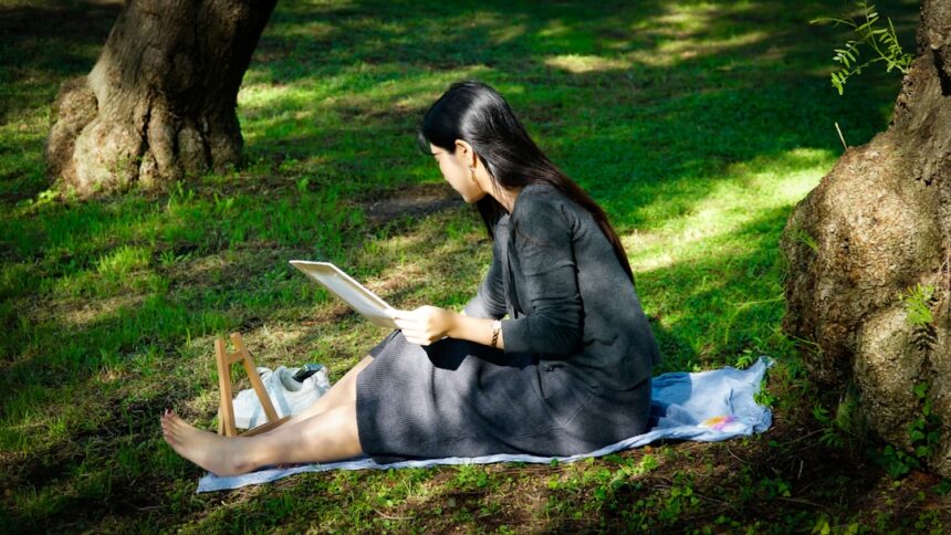 Woman reading tablet on blanket in grassy park.