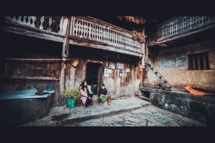 Two people sit outside an old wooden house.