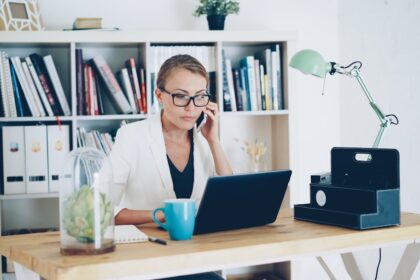 Woman on phone at desk with laptop and books.