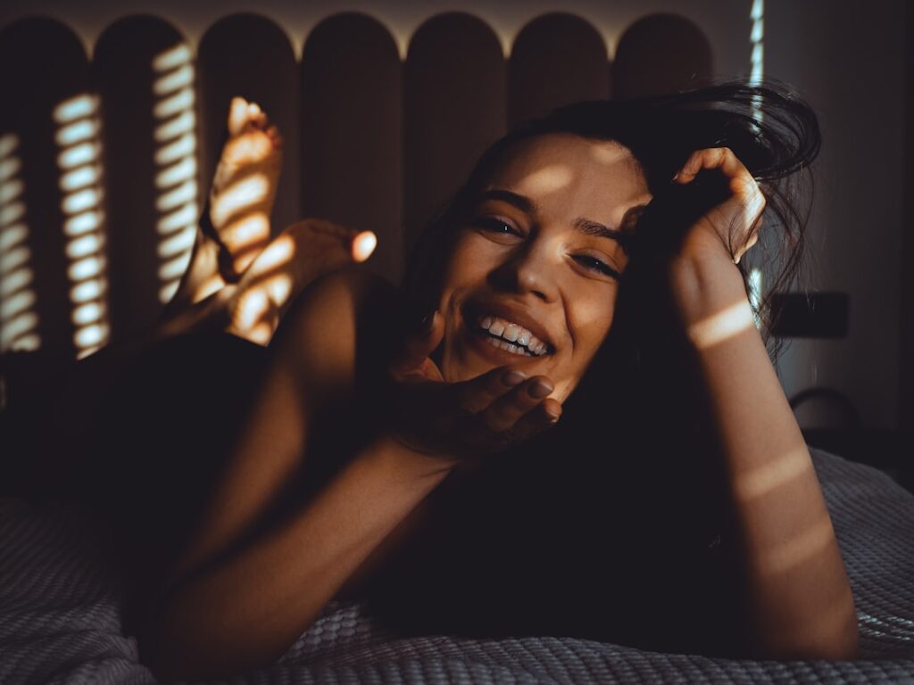 A smiling woman lies on a bed with striped shadows.